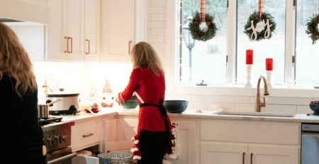 a woman puts bowls away into a kitchen drawer. The kitchen is white and contemporary. there are three christmas wreaths hanging in the window.